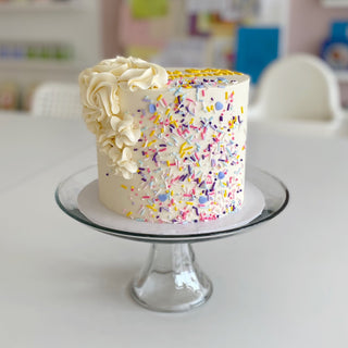 White cake with white rosettes and colourful sprinkles splashed up the side. Cake is on a clear cake stand on a white table. 