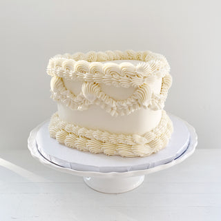 White lambeth heart cake with vintage piping viewed from the side. Cake is on a frilled white ceramic cake stand. 