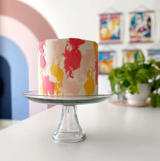 White cake with abstract patches of peach, yellow and hot pink. Cake is on a clear cake stand. 